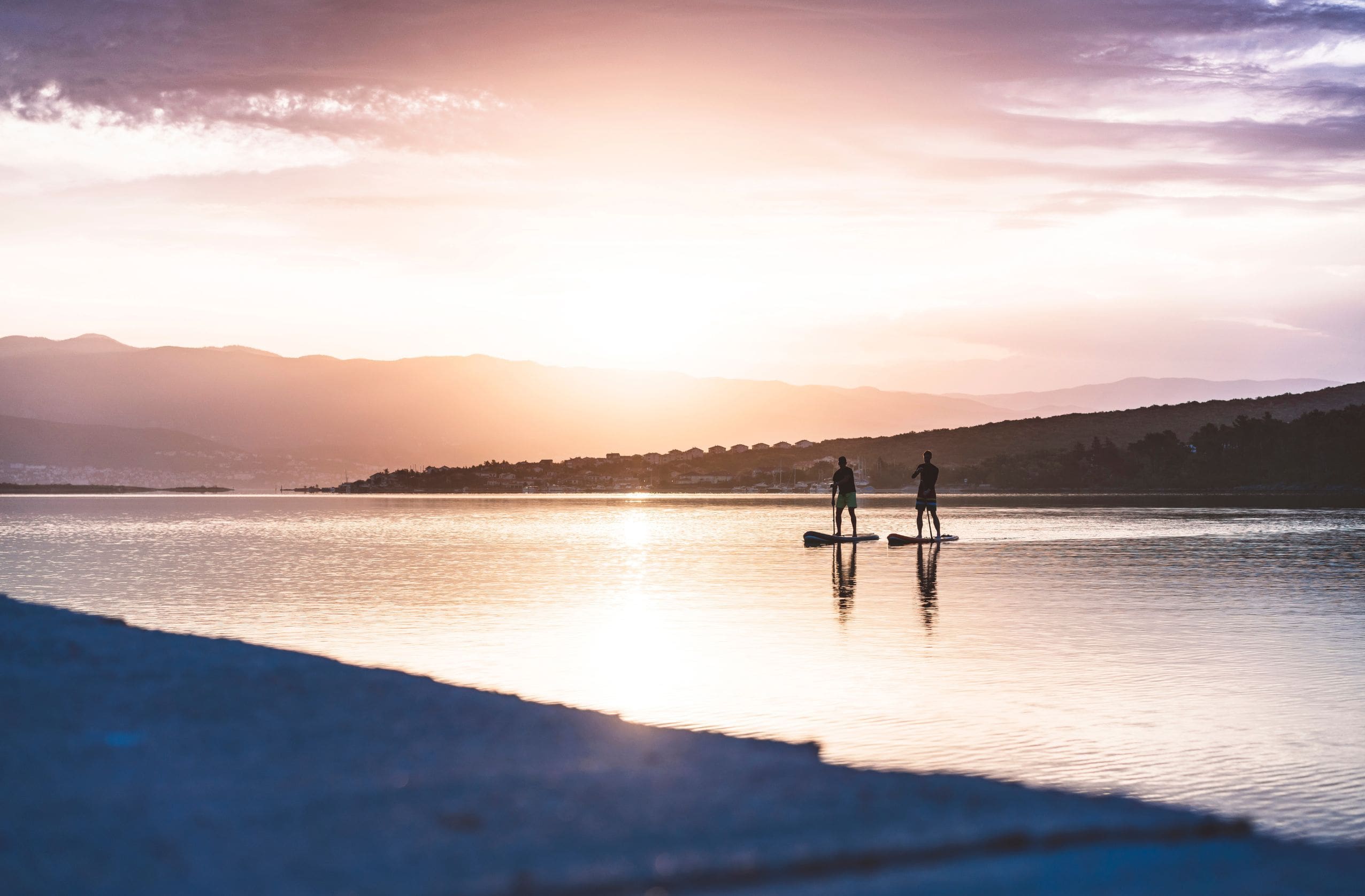 Two people paddleboarding on a calm lake at sunrise, capturing the peaceful vibe of SUP adventures and the ideal setting for waterproof Bluetooth speakers.